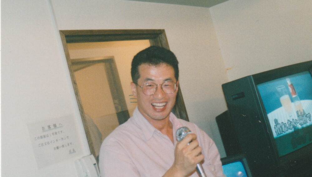 カラオケボックスで、銀色のマイクを握って笑顔の小野先生